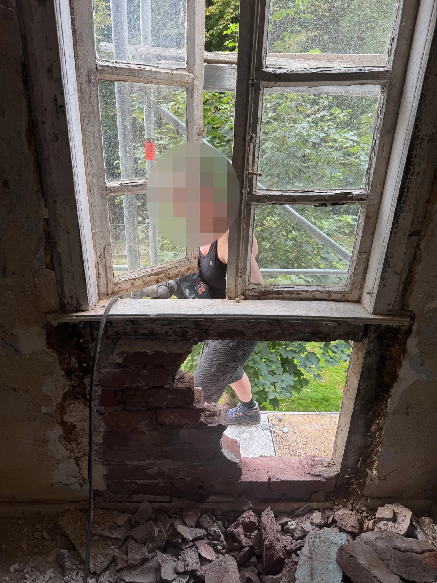 Blick von innen durch ein Fenster: Draußen auf dem Gerüst arbeitet eine Person, darunter klafft ein Loch in der Ziegelausmauerung, davor liegt Schutt.