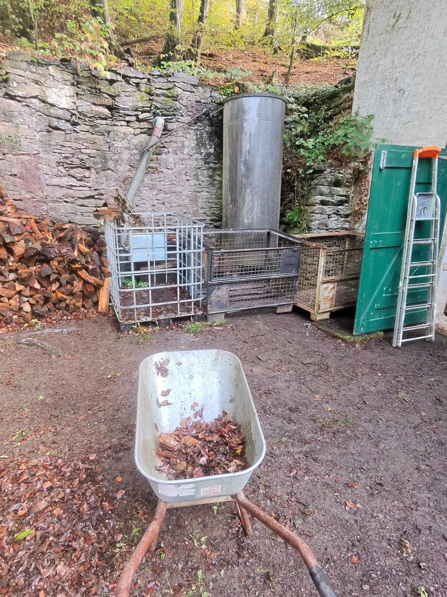 Eine Schubkarre mit Laub vor ordentlich aufgereihten Gitterboxen, daneben ein Holzstapel und eine Leiter am grünen Tor.