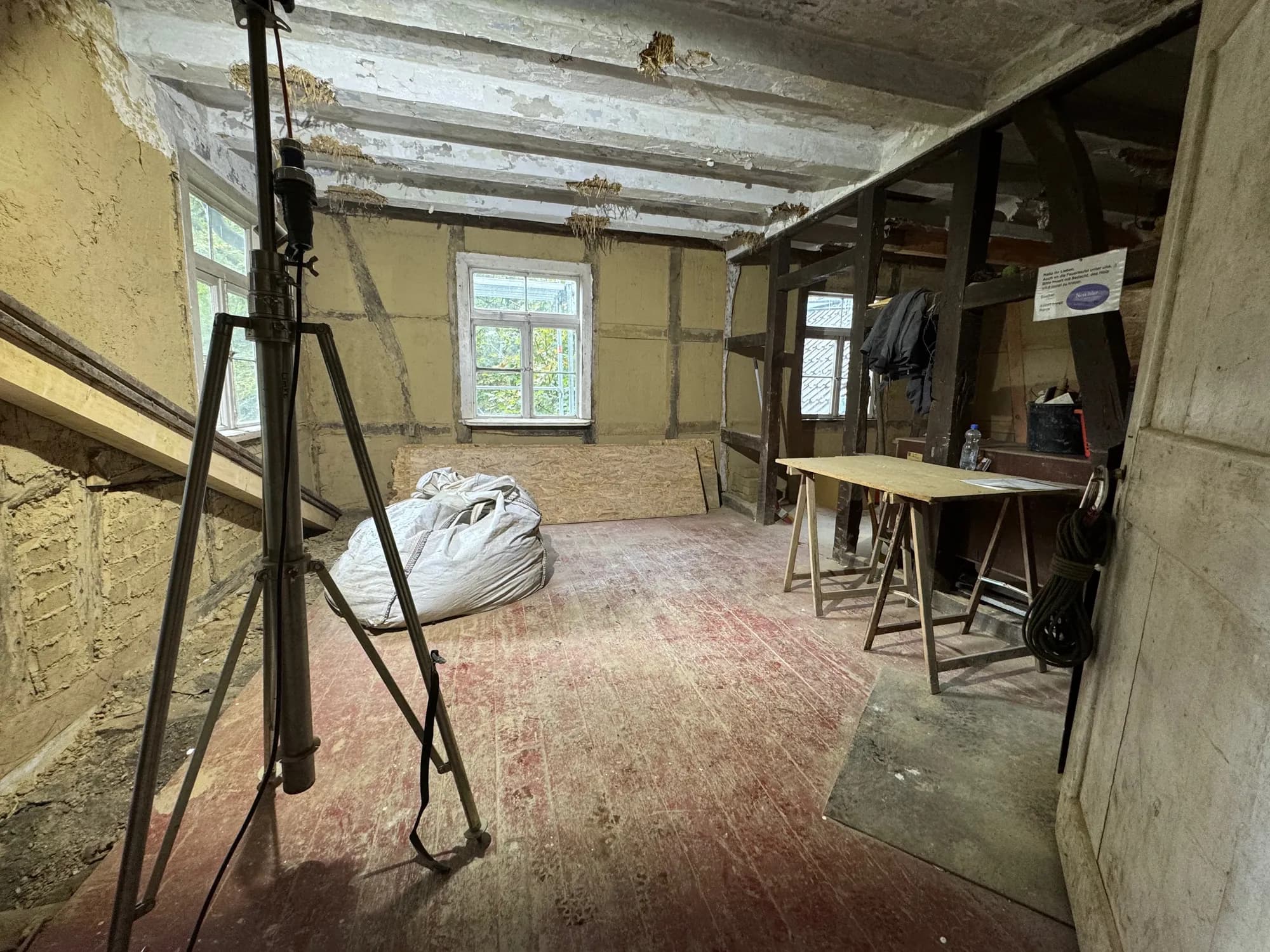 Aufgeräumter Raum im Wohnhaus mit Lehmwänden, gefegtem Dielenboden, Arbeitstisch auf Böcken und einem Stativ im Vordergrund.